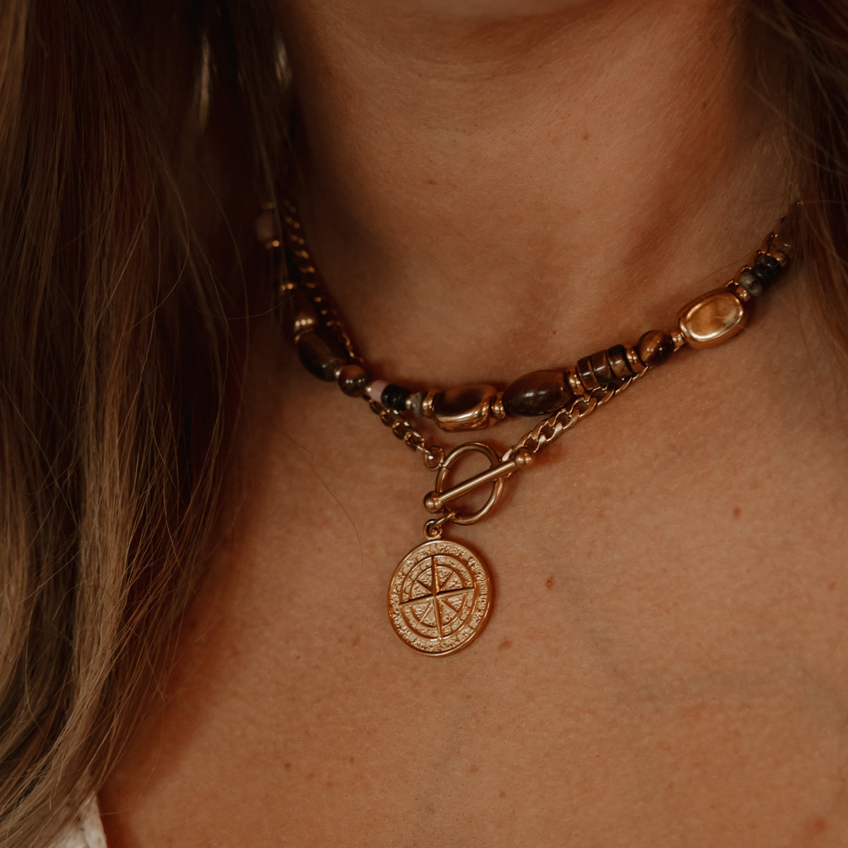  The necklace features a dark brown beaded top layer, a gold-toned chain middle layer with a toggle clasp, and a circular gold compass pendant charm.